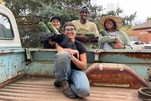 A young Asian women kneels in the rusty bed of a pickup truck, posing with three people standing people her, each holding large green gourds.