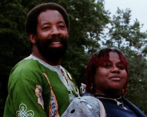 A man with short dark Afro and bears and a woman with red-dyed locs pose and smile.
