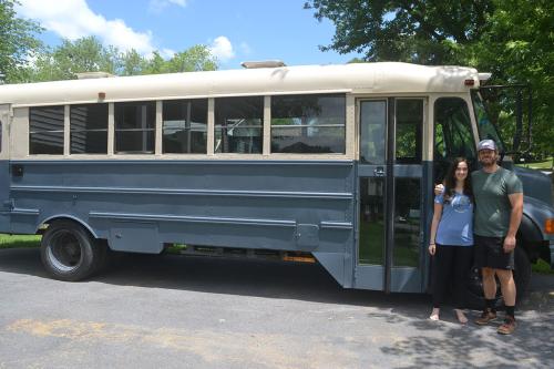 A man and woman pose in front of a school bus that has been painted blue and white.
