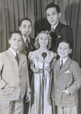 Five children, four boys and a girl, pose smiling in front of a microphone in formal wear.