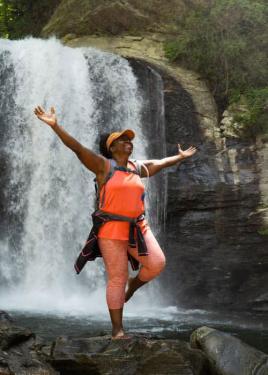 A woman wearing orange visor, tank top, and leggins and hydration backpack poses barefoot on a rock in front of a waterfall, balancing on one foot with hands outstretched.
