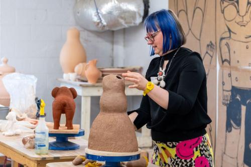 A woman with chin-length blue hair and a woven skirt with neon pink happy faces works on a clay sculpture in a workshop.