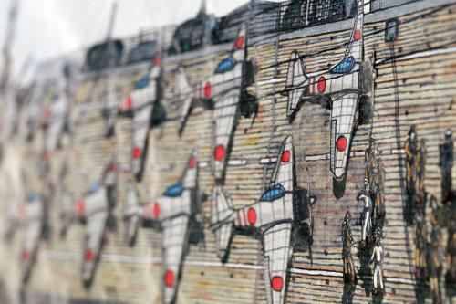 Closeup, at an angle, of colored pencil drawings of jets lined up on the deck of a ship.
