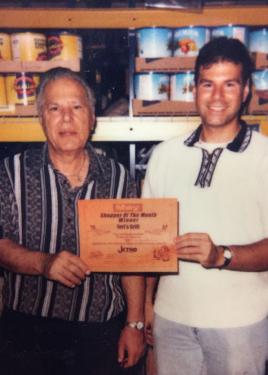 Two  men, father and son, pose both holding a document that reads “Shopper of the Monther Winner: Jeri’s Grill. Bulk cans of food products on shelves behind them.