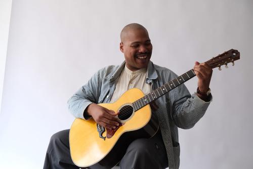 Jerron laughs as he plucks an acoustic guitar. The edge of the white backdrop is visible in the corner.