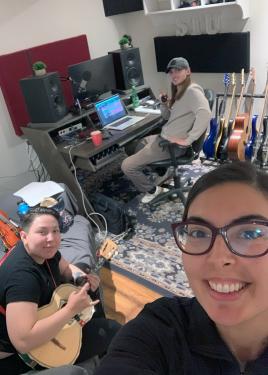 Selfie of Jessie, a Latina woman, with a person on a small guitar-like instrument and a person at a desk with laptop behind her.