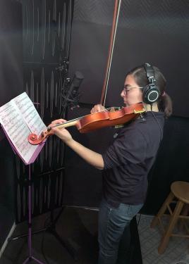 Jessie plays violin in front of a microphone and music stand, wearing headphones, in a black room.