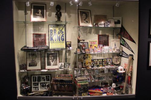 Museum display case showing photos and paraphernalia of African American entertainers and athletes. Notable items: BB King poster, Duke Ellington record, figure of Muhammad Ali.