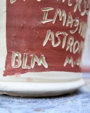 Closeup on a white ceramic jug, possibly before it has been glazed and fired. White carved letters on a square red background read BLM.
