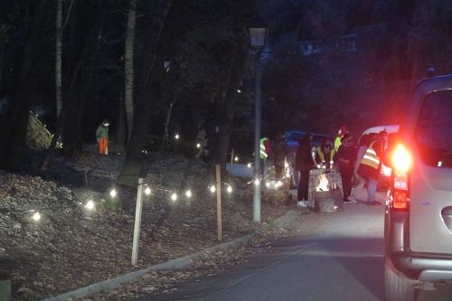 View of a line of cars at dusk on a one-lane road. A group of people speak with the driver of one car.