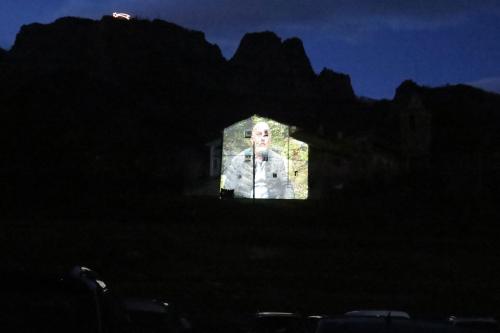 View from the parked cars: a projection of a man on the side of a church building. In the distance, an illuminated shooting star on the ridge of a mountain.