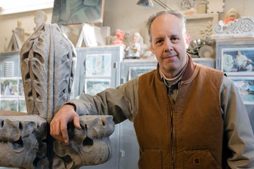 Joe smiles, resting one arm on a carved stone piece inside the masonry workshop, surrounded by tools and cabinetry.