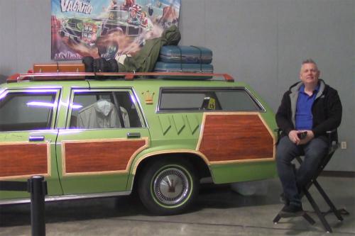 A man sits next to a green, wood-paneled station wagon, topped with luggage on the roof rack, with a poster for National Lampoon’s Vacation hanging on the wall behind it.