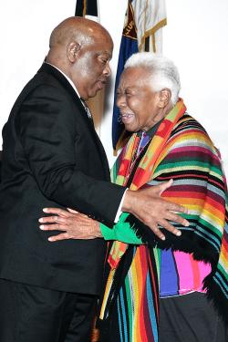 An older man in a suit and older woman in a colorful striped shawl embrace.