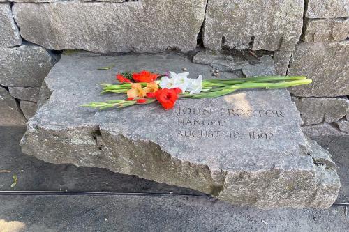 Gray flagstone jutting out from a stone wall, with words etched in: John Proctor, hanged August 19, 1692. Above the lettering, white and orange cut flowers.