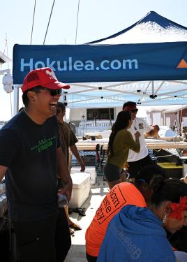 A young man wearing black shirt and pants, red trucker hat, and sunglasses laughs, standing above others seated. Behind them, a popup tent with text on the side: Hokelea.com.