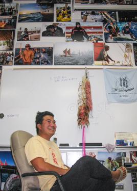A young man sitting in an office chair laughs. Behind him are a whiteboard and large photographs of a crew at sea covering every inch of wall.