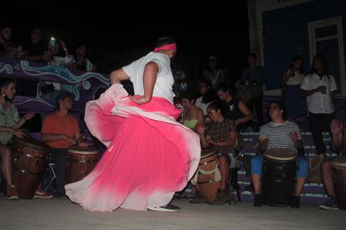 A man in a long, flowing hot pink skirt dances in front of a line of seated drummers. He is illuminated, but the scene is otherwise in the dark of night.