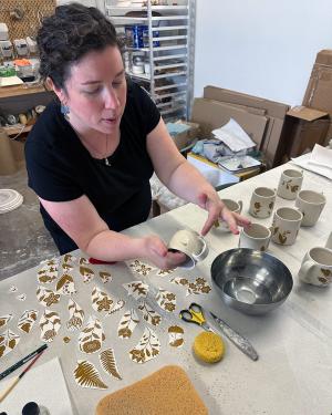 A woman with curly brown hair holds a small mug with one hand, and points to the bottom with the other. On the table in front of her are several white cups with floral designs in brown, and cutouts of more of the floral designs, like stickers.
