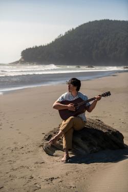 A man plays acoustic guitar, sitting on a rock on an oceanside beach.
