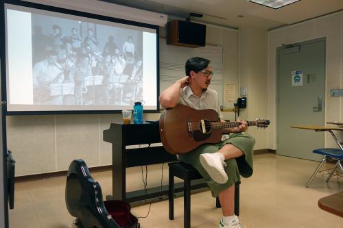 Julian, an Asian man with glasses and goatee, sits at the front of a classroom with an acoustic guitar and a black-and-white photo of an orchestra on screen behind him.