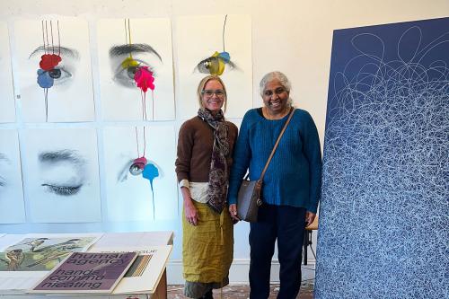 Two women pose among paintings of eyes on the wall and an abstract painting of white swirly lines on dark blue.