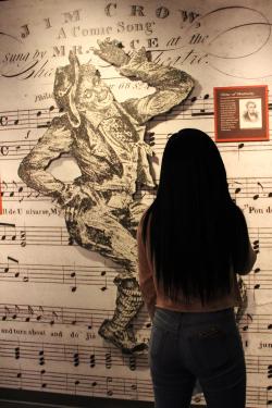 A woman stands in front of a museum display, with blown-up sheet music of the song “Jump Jim Crow” and a cartoon of the title character.