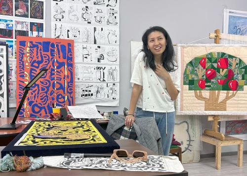A woman speaks in an art studio. Behind her are abstract illustrations and a quilt of a stylized apple tree.