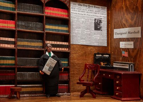Close-up of a diorama showing Thurgood Marshall, wearing glasses and a suit, in an office with tall book shelves. On the wall, a New York Times article with the headline: High court bands school segregation.