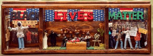 Close-up of a wide diorama in three sections, with a banner across the back reading Black Lives Matter. On the left, Scrabble tiles spell out Trayvon Martin. A figure in a white hoodie holding a bag of Skittles raises us his hands. In the center, a family