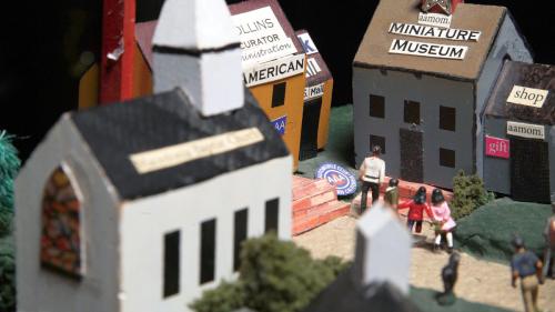 Close-up of a diorama of a city square, with a church building out of focus in the foreground, and a building labels Miniature Museum in focus. A miniature family walks up to its tiled stairs.