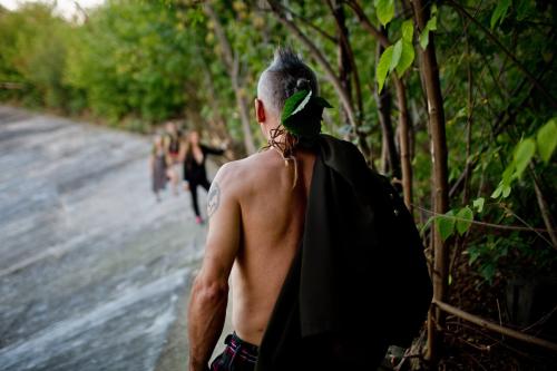 From behind, a shirtless man walks along the edge of a sloped, paved surface, with dense trees lining the edge. He heads toward other people out of focus in the distance.