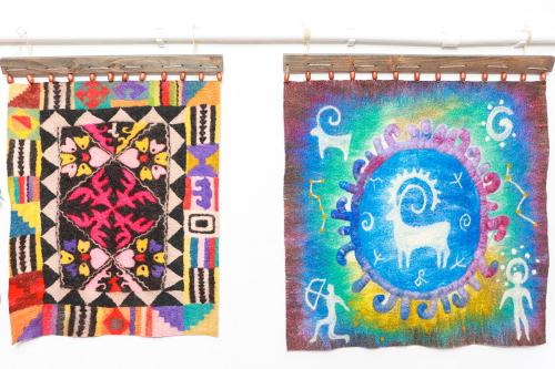 Two felt textiles hanging. On the left, a mandala design with floral and geometric shapes in bright colors. On the right, a horned animal like a deer in the center of a blue circle, with rays that radiate outward like the sun, toward four figures in the c