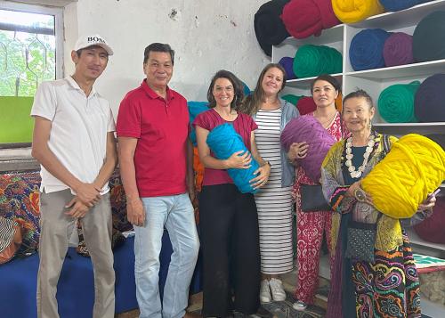 Six people pose in a studio. Three of them hold huge bundles of brightly dyed wool in blue, purple, and yellow. Other bundles line the shelves behind them.