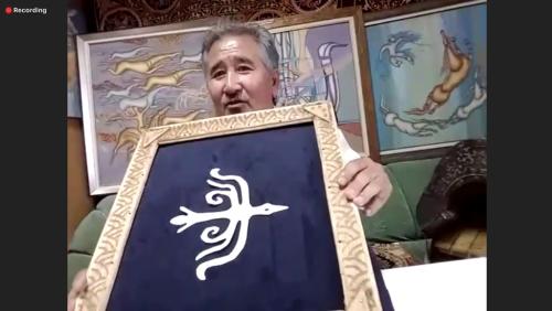 Zoom screenshot showing just the man from the previous image, holding up a dark blue framed textile with the image of a white stylistic bird.