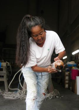 A woman with long dark hair in a ponytail and ripped jeans leans over, working with a piece of wood.