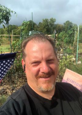 A man stands in front of a green garden, the stars of an American flag visible on one side.