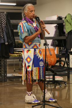 Man with long braids and blue and white regalia plays a long wooden flute.