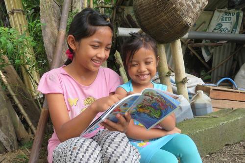 Two young girls read a comic book together, giggling.