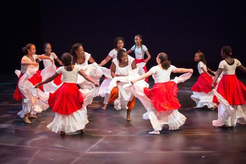 On a theater stage, ten child and adult dancers dressed in white and red skirts and white tank tops form a loose circle.