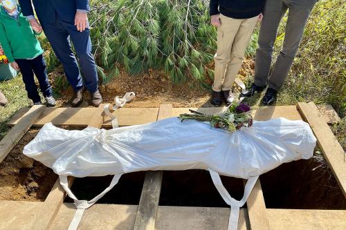 A body wrapped in white fabric, laid on wooden planks over an open grave. A bouquet of flowers is on its chest.
