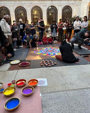 The same scene later in the day: the two women have completed a central diamond-shaped colorful design, and now add white embellishments to fill out the corners of their square platform. In the foreground are shallow golden bowls of bold pigments; red, or