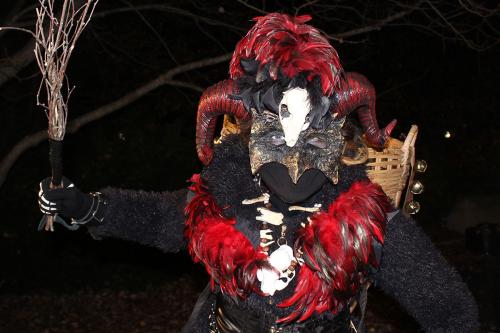 Person dressed in a Krampus costume, with a red feather boa and horned headdress. It holds a bundle of sticks menacingly, as if about to strike.