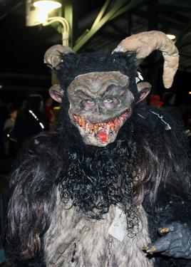 A person in a masked monster suit, complete with blood-stained teeth, brown hairy body, curved horns, and a ghoulish face mask, poses among a crowd of people.