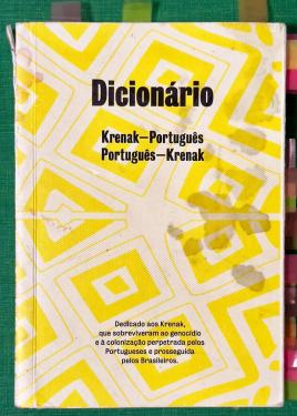 Cover of a book with white and yellow geometric patterns and  black text: Dicionário. Krenak-Português, Português-Krenak. The cover is well worn, with dark water stains. The spine is peeling slightly.