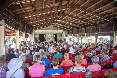 Frmo the back of an open-air barn, a seated crowd faces a quilt hanging above the stage in front, suspended between two men on ladders.