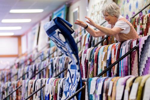 A woman throws a quilt off a rack. Other are neatly lined up in three rows.