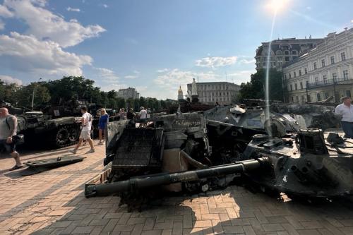 In a brick-lined city square, people walk among destroyed tanks and artillery. The sun shines brightly overhead.