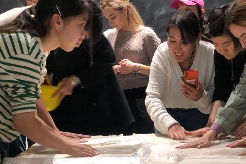 Women gather around a table, touching samples of white fabric laid flat.