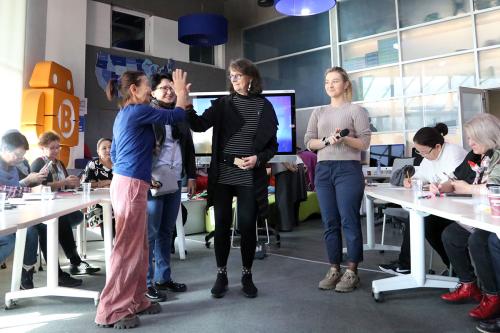 Two women high-five as two others stand next to them. On either side, women sit at worktables.